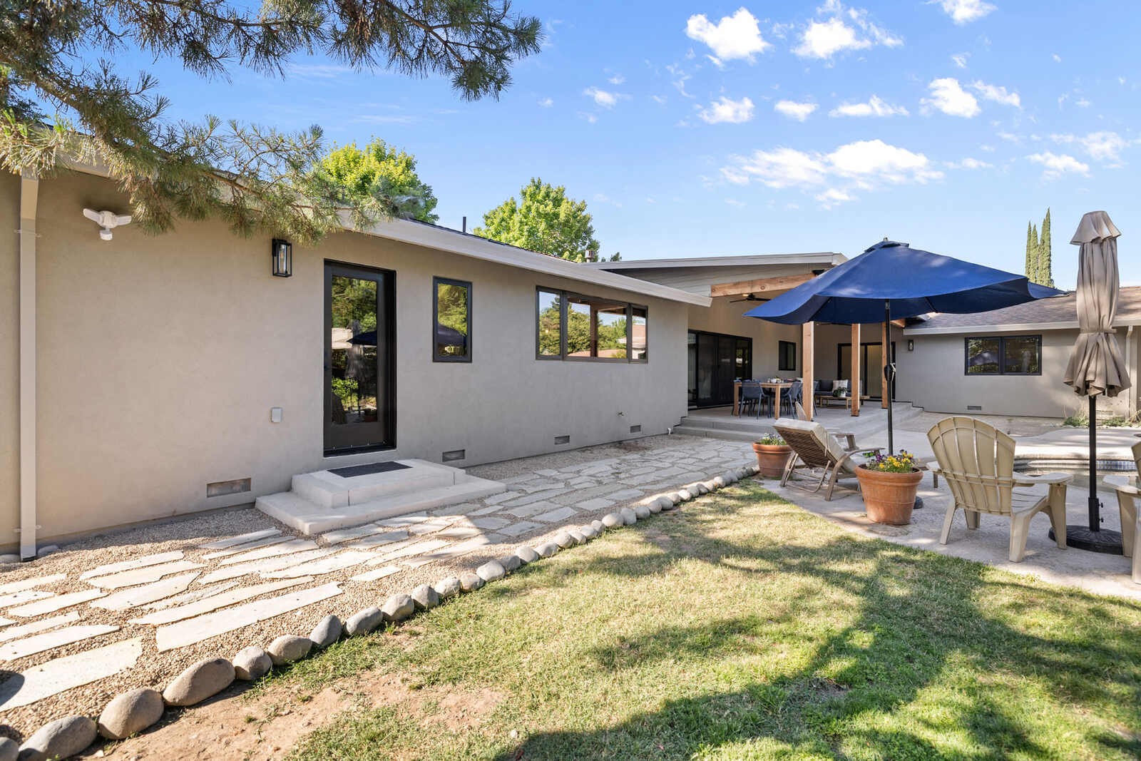 Backyard patio with stone pavers, lawn, and outdoor seating with blue umbrella behind a modern single-story home in Sacramento Backyard patio with stone pavers, lawn, and outdoor seating with blue umbrella behind a modern single-story home in Sacramento