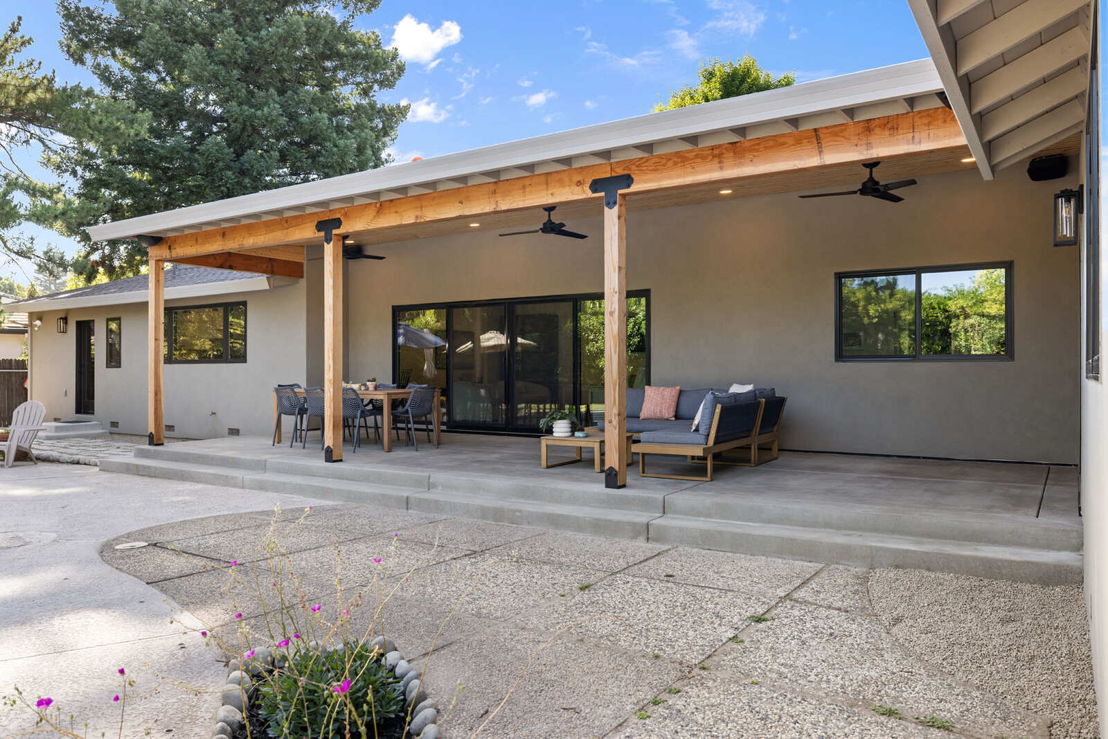 Covered backyard patio with wood posts, ceiling fans, seating area, and sliding glass doors at a modern single-story home in Sacramento